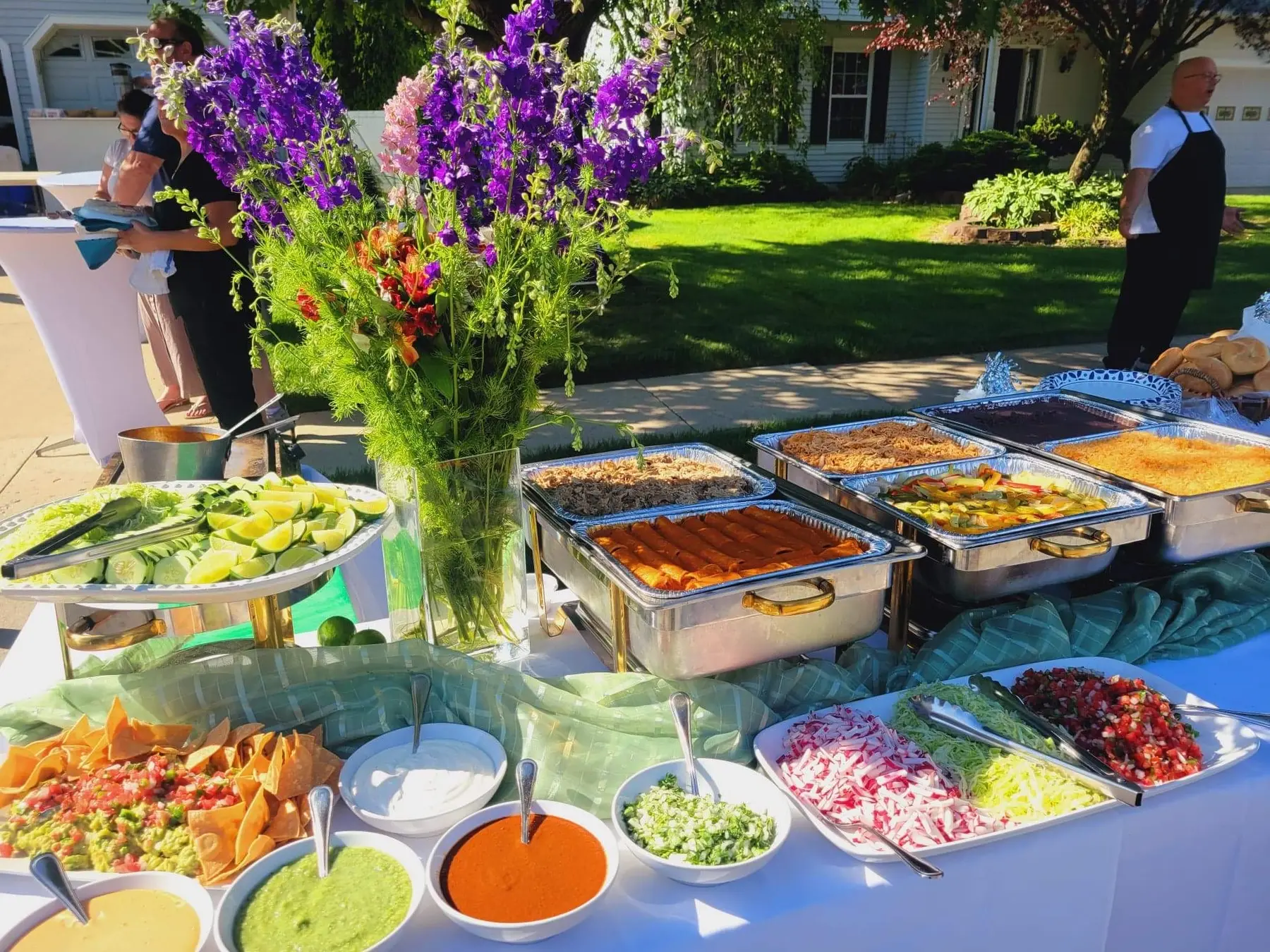 An outdoor catering spread of trays beside purple flowers