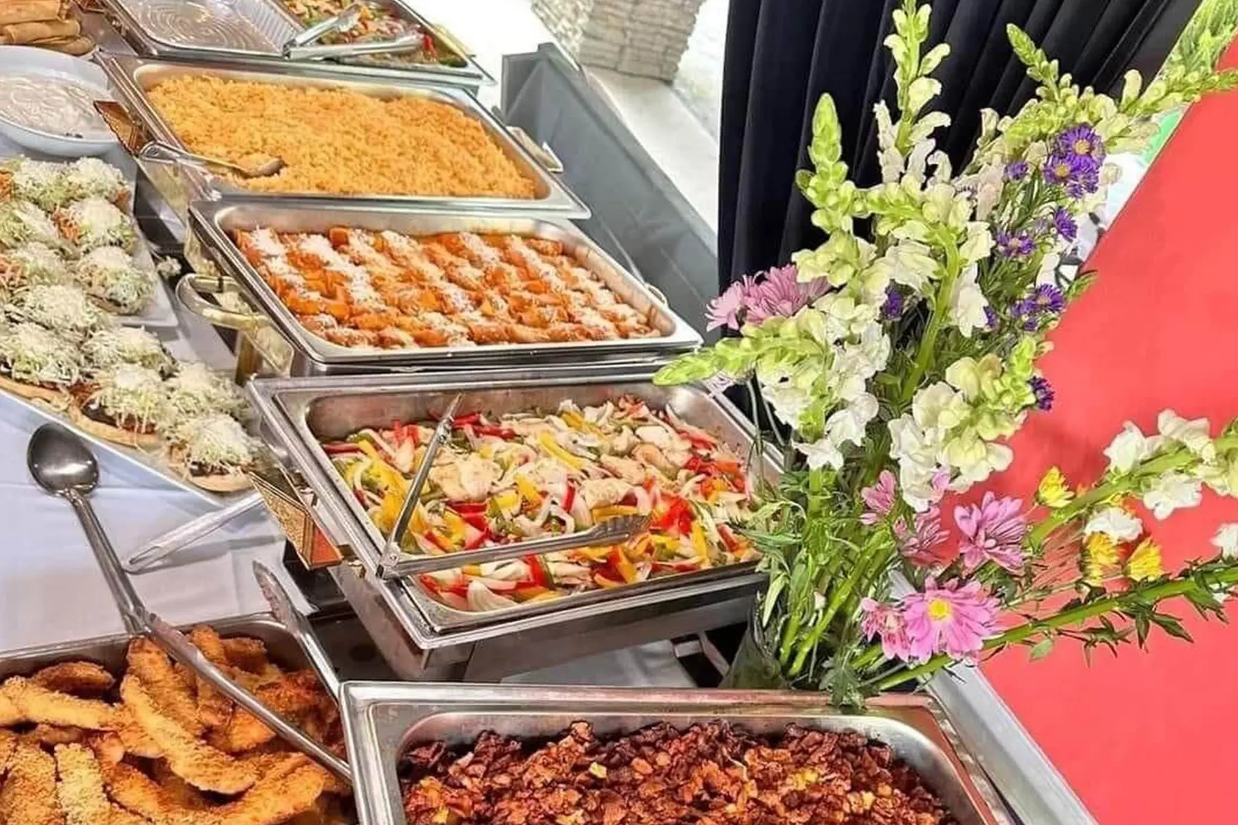 Catering trays of rice, beans and grilled meats beside fresh flowers