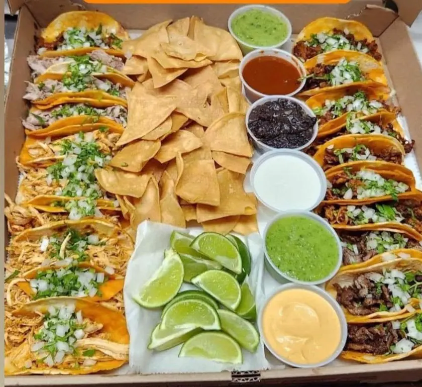 Twenty tacos with cilantro and onion beside chips, limes and six salsas
